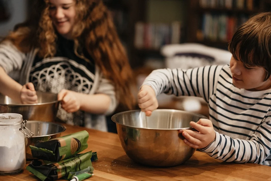 A young child wearing a small apron and chef hat rolling out dough on a flour-dusted kitchen counter beside a parent, with colorful silicone baking tools nearby